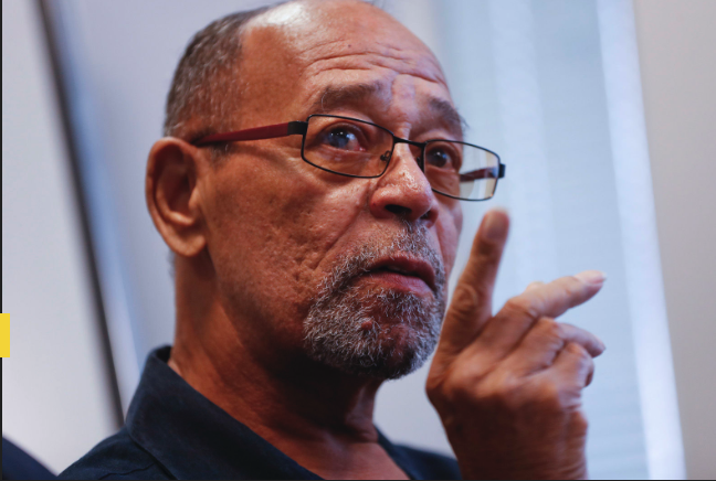 A close-up photograph of an older man with glasses and a goatee, pointing his finger while speaking.