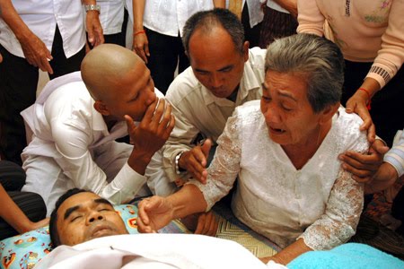 A photograph showing a group of Cambodian people mourning over a deceased man. An elderly woman in a white lace shirt is crying and reaching out to the body, while others around her offer support.