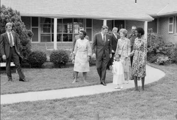 A black and white historical photograph showing President Ronald Reagan and First Lady Nancy Reagan walking on a sidewalk in front of a brick house, accompanied by a young girl and several other people, including Secret Service agents.
