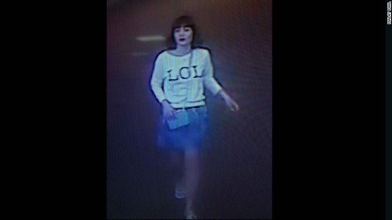 A low-resolution CCTV screenshot of a woman with dark hair and bangs, wearing a white long-sleeved shirt with the word 'LOL' printed on the front.
