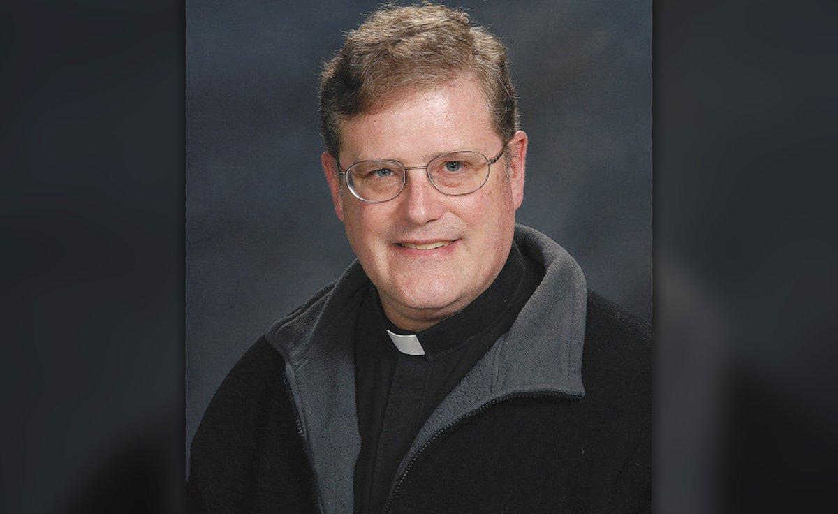 A portrait of a middle-aged Caucasian man with reddish-brown hair and glasses, wearing a Catholic priest's clerical collar and a black fleece jacket.
