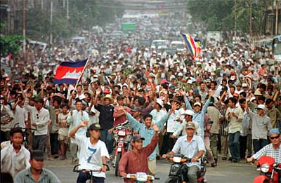 A large crowd of Cambodian people marching down a city street, some on motorbikes, waving and holding a Cambodian flag.