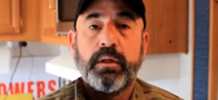 A close-up photograph of a man with a full grey and black beard wearing a black baseball cap and a tan shirt.