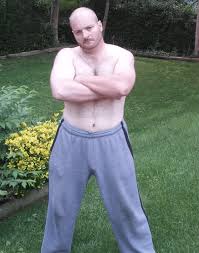 A shirtless Caucasian man with a shaved head and a mustache, standing with arms crossed in a grassy yard, wearing grey sweatpants.
