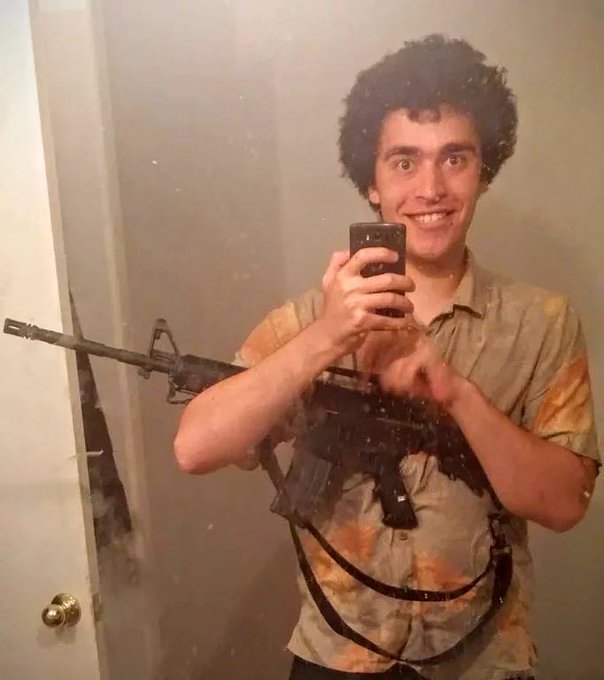 A mirror selfie of a young man with dark curly hair, smiling and holding an assault-style rifle across his chest while taking the photo with a smartphone.