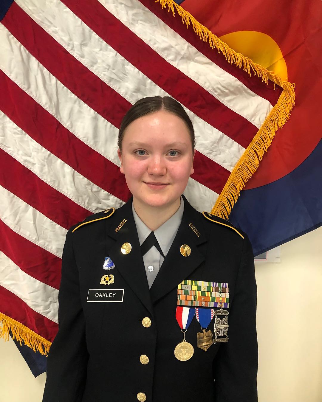 A portrait of a young woman in a JROTC uniform with various medals and ribbons, standing in front of an American flag and a Colorado state flag.