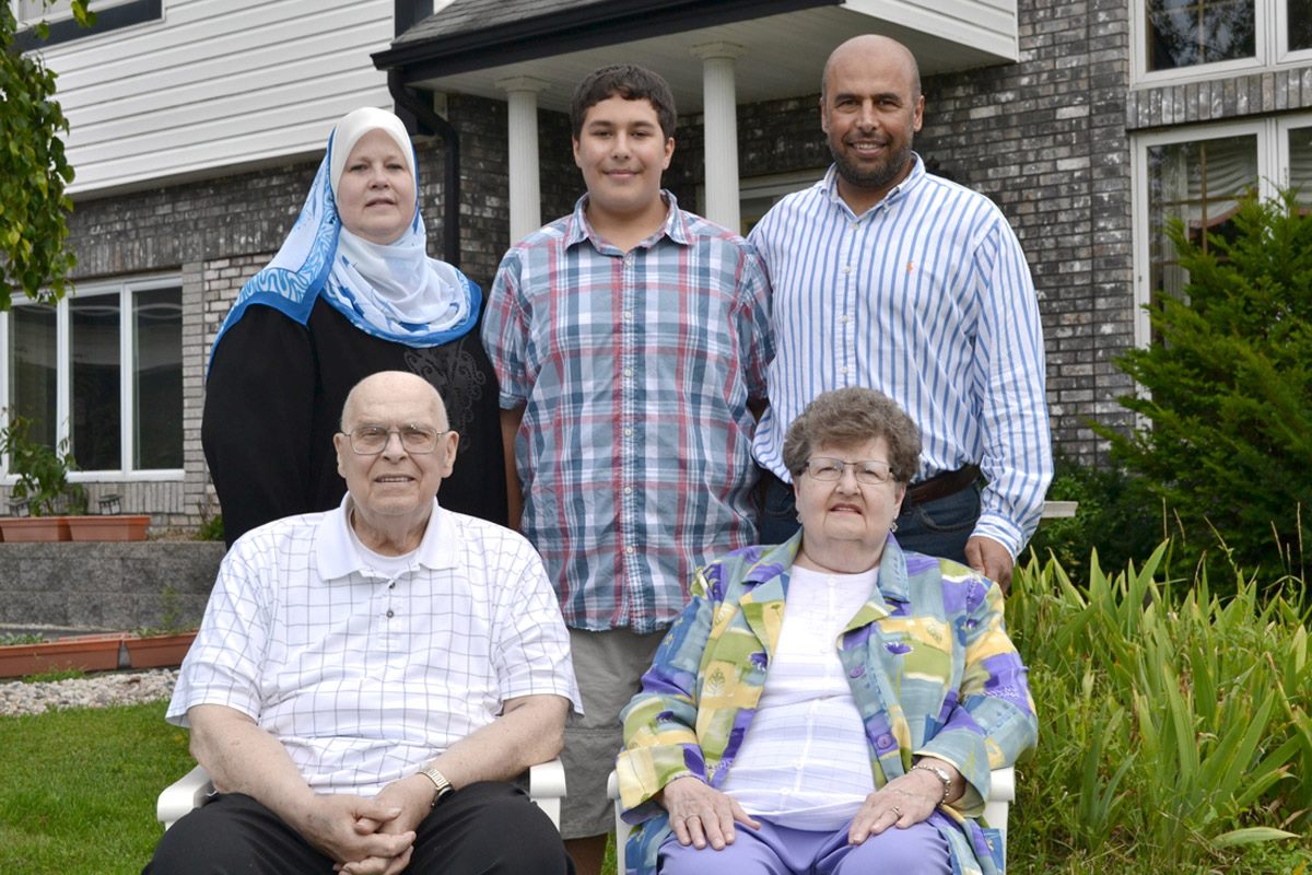 A family portrait of five people in front of a house. Two elderly individuals are seated in the foreground, while three younger adults stand behind them. One woman wears a blue and white hijab.
