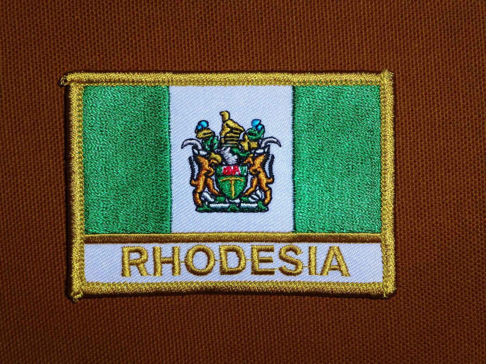 An embroidered patch of the Rhodesian flag with the word 'RHODESIA' written below it in gold letters, set against a brown fabric background.