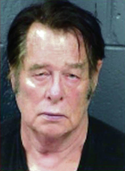 A mugshot of an older Caucasian man with dark hair and sideburns, wearing a black shirt against a dark textured background.