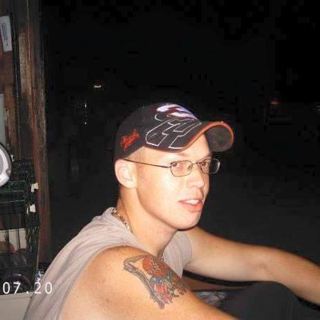 A photograph of a young Caucasian man wearing a black baseball cap with red and white embroidery, thin-rimmed glasses, and a grey sleeveless shirt. He has a colorful tattoo on his right shoulder and is looking off-camera to the right.