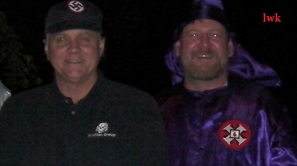 A photograph of two men at night. The man on the left wears a black cap with a swastika and a black shirt with a skull logo. The man on the right wears a purple Ku Klux Klan robe with a hood and a red cross patch.