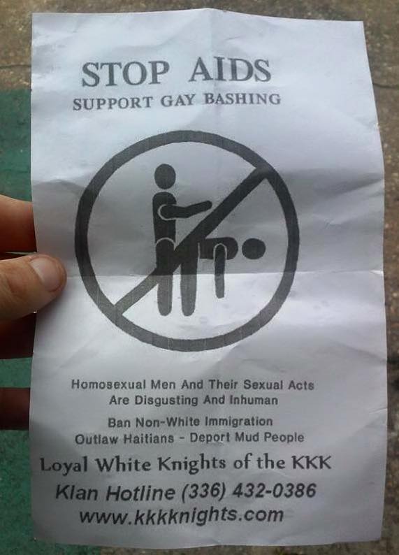A photograph of a hand holding a crumpled white flyer containing hate speech and extremist propaganda from the Loyal White Knights of the KKK. The flyer includes a graphic against homosexuality and text calling for violence and the deportation of non-white people.