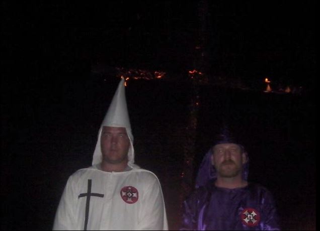Two men in Ku Klux Klan robes, one in white and one in purple, standing in front of a burning cross at night.