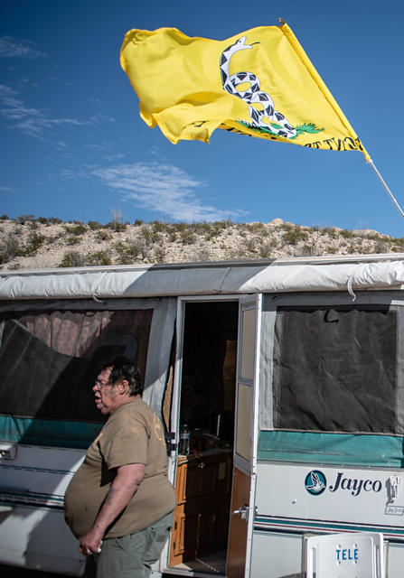 A man with dark hair and a heavy build stands outside a Jayco camper in a desert setting, with a yellow Gadsden flag flying above.