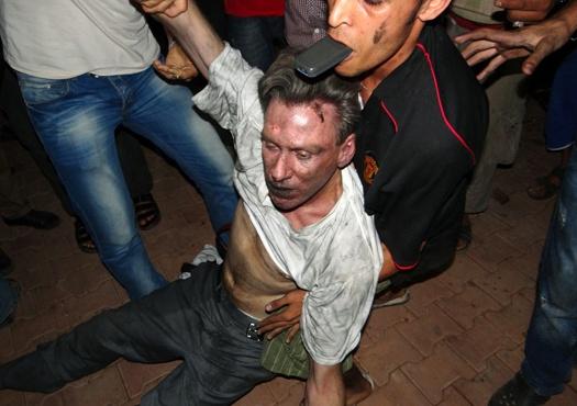 A photograph showing a group of men carrying an unconscious or injured Caucasian man through a crowd at night. The man being carried has dark hair and is wearing a white shirt. One of the men carrying him has a mobile phone in his mouth.