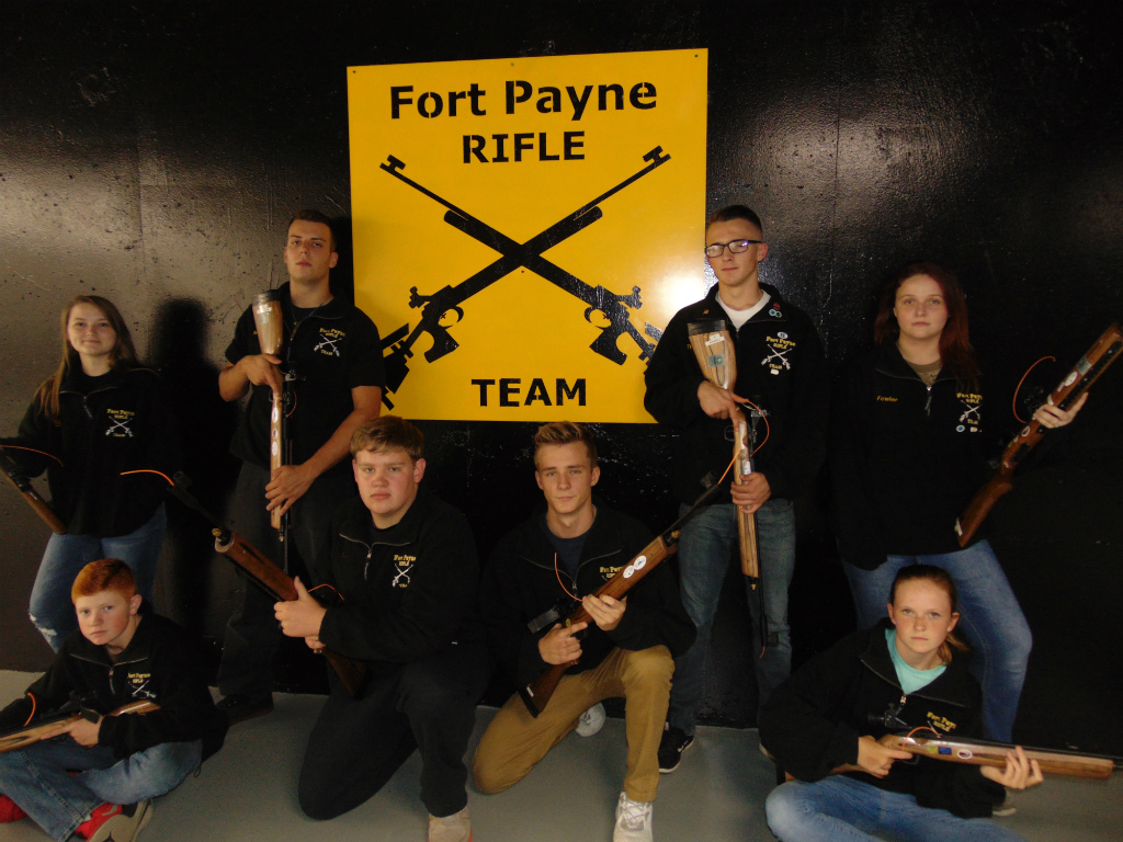 A group of eight teenagers posing with rifles in front of a yellow sign that reads 'Fort Payne RIFLE TEAM'.