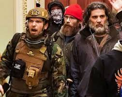 A photograph showing several men inside a building during a protest or riot. One man in the foreground wears a tan tactical helmet and vest over camouflage clothing, appearing to shout. Behind him is a man with a thick brown beard wearing a red beanie and glasses. To the right is another man with long dark hair and a grey-flecked beard.