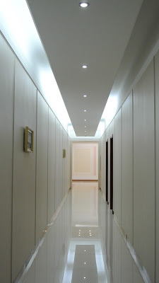A long, narrow, brightly lit hallway with white paneled walls and a highly reflective floor. Small framed pictures hang on the left wall, and dark doorways are visible on the right.