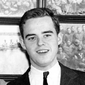 A black and white portrait of a young Caucasian man with dark wavy hair, wearing a suit and tie, standing in front of framed sports photographs.