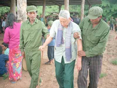 A photograph showing an elderly Asian man with white hair, identified as Pol Pot, being assisted by two men in green military uniforms in a rural setting.