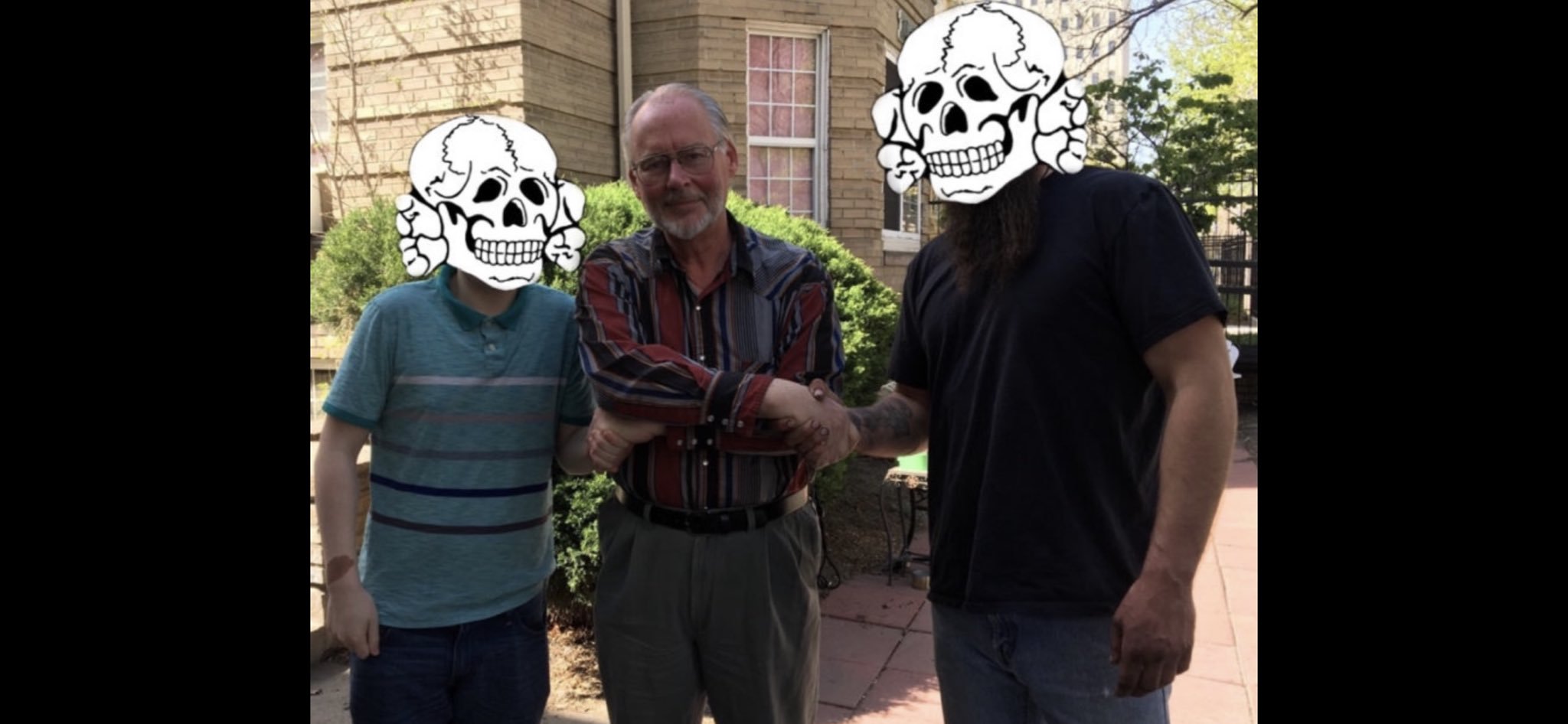 A photograph of three men standing outside a building. The man in the center is an older Caucasian male with grey hair, glasses, and a beard, wearing a striped shirt. The faces of the men on either side are obscured by cartoon skull illustrations. The man on the right has a long dark beard.