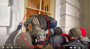A video screenshot showing a group of rioters at the US Capitol on January 6th, with one individual in tactical gear highlighted by a red circle as they attempt to enter through a window.