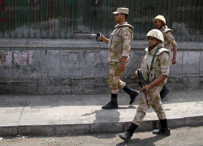 Three soldiers in desert camouflage uniforms walking along a sidewalk next to a weathered wall. The soldier in the center is holding a handheld device with a long, thin antenna, while the soldier in the foreground carries a rifle.