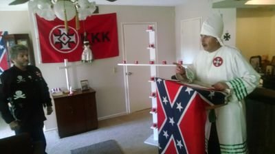 A photograph showing a man in a white Ku Klux Klan robe and hood speaking at a podium draped with a Confederate flag. Another man in a black jacket with skull and crossbones patches stands nearby. A red KKK flag and a white cross are visible in the background.
