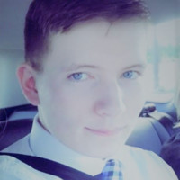 A close-up photograph of a young Caucasian male with short brown hair and blue eyes, wearing a white dress shirt and a blue patterned tie, sitting in a vehicle.