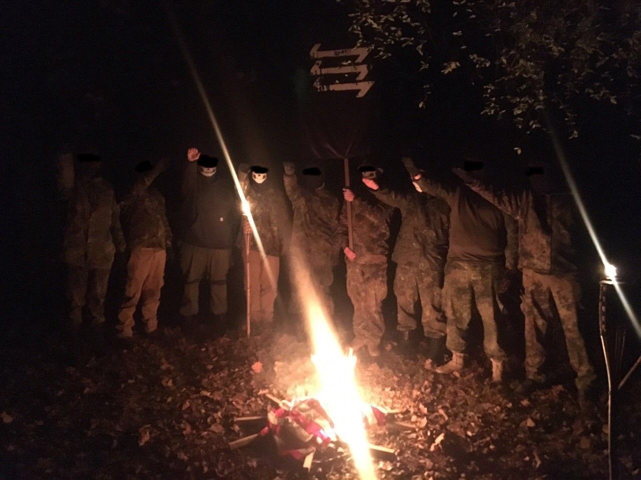 A group of individuals in camouflage and masks standing around a bonfire at night, performing a Nazi salute. A flag with a white symbol on a black background is held in the center.