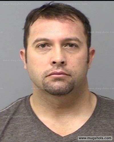 A mugshot of a Caucasian man with short dark hair and a goatee, wearing a grey t-shirt. The image contains watermarks from mugshots.com.