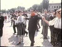 A historical photograph showing a man in a dark suit and glasses dancing in a circle with people wearing traditional folk costumes in an outdoor plaza.