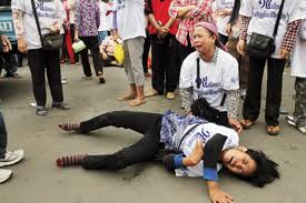 A photograph showing a woman lying on the ground in distress during a protest in Cambodia, with another woman kneeling beside her in grief. They are surrounded by bystanders wearing matching white t-shirts.