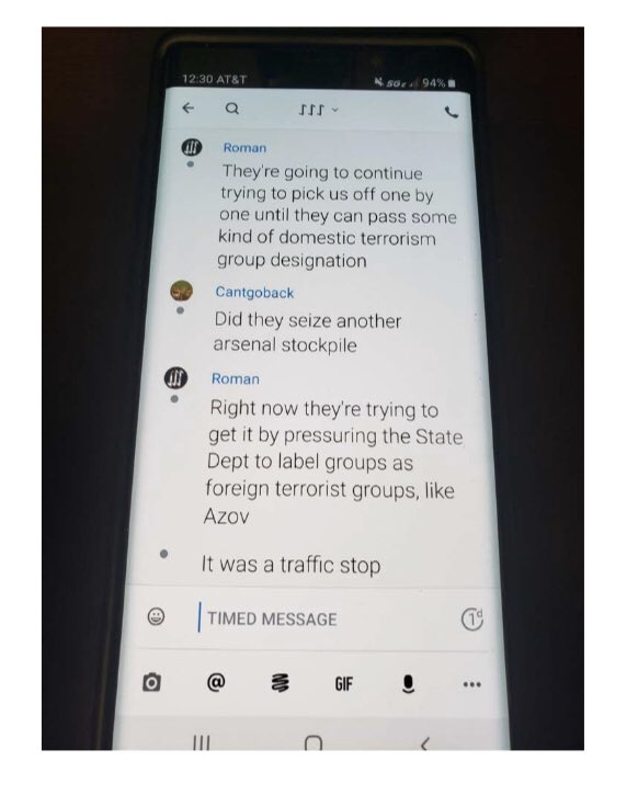 A photograph of a smartphone screen showing a chat log. The messages from 'Roman' and 'Cantgoback' discuss government efforts to designate domestic groups as terrorist organizations and mention the Azov movement.