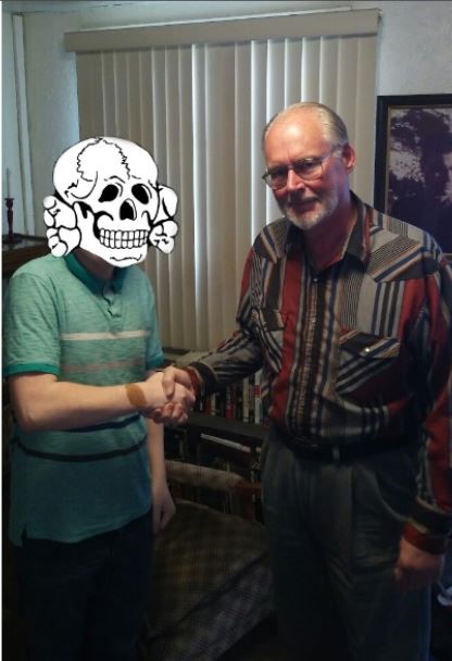 A photograph of two men shaking hands indoors. The man on the left has his face obscured by a cartoon skull graphic and is wearing a teal polo shirt. The man on the right is an older Caucasian male with glasses, a grey beard, and a full head of thinning hair, wearing a multi-colored patterned button-down shirt.