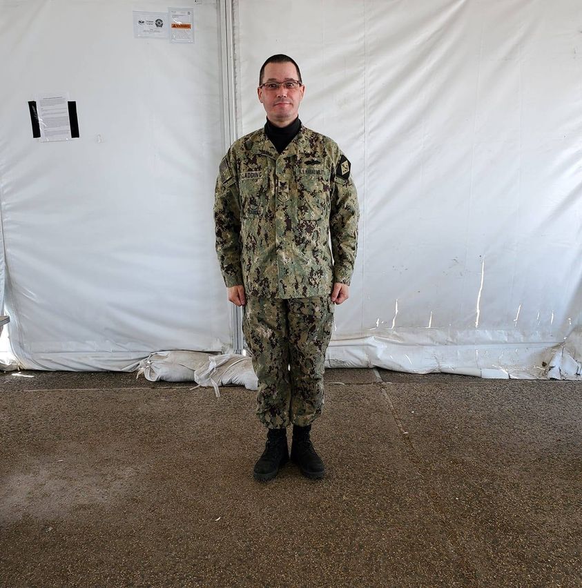 A man in a New York Naval Militia camouflage uniform standing at attention in front of a white tent wall. He is wearing glasses and has short dark hair.