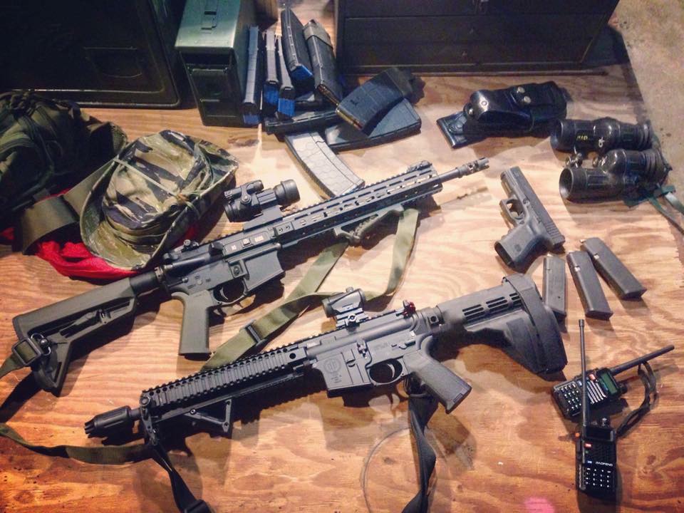 A collection of tactical gear and firearms laid out on a wooden table, including two AR-15 style rifles, a handgun, multiple magazines, binoculars, a radio, and a camouflage hat.
