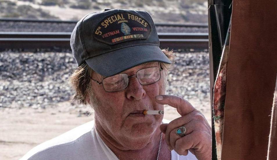 A close-up photograph of Larry Mitchell Hopkins, an older man with glasses and a '5th Special Forces Vietnam Veteran' baseball cap, smoking a cigarette outdoors near railroad tracks.