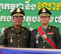 Two Cambodian military officers in formal uniform standing against a green backdrop with Khmer text.