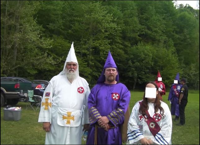 A photograph of several individuals in Ku Klux Klan robes standing in a field. One man wears a white robe and has a long white beard; another wears a purple robe and has a goatee. A woman in a white robe has her face censored. Trees and vehicles are in the background.
