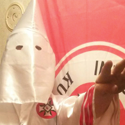 A person wearing a white Ku Klux Klan hood and robe with a red patch, standing in front of a red and white flag.
