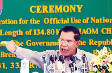 A photograph of Cambodian Prime Minister Hun Sen speaking forcefully at a podium during a ceremony, pointing his finger while standing in front of a green backdrop with yellow lettering.