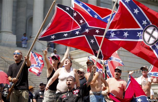 A group of white supremacists holding Confederate and KKK flags on the steps of a building, shouting and gesturing.