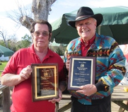 Two men standing outdoors holding award plaques. One man wears a red shirt and glasses, while the other wears a black cowboy hat and a patterned jacket.