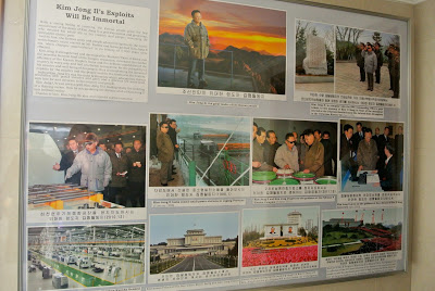 A propaganda display board titled 'Kim Jong Il's Exploits Will Be Immortal' featuring several photographs of the former North Korean leader Kim Jong Il in various settings, accompanied by text in English and Korean.
