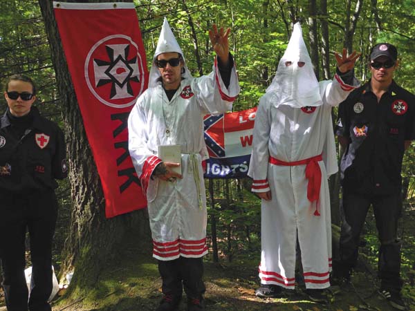 A photograph of four individuals associated with the KKK in a wooded setting. Two are wearing white robes and pointed hoods, while two others wear black shirts with KKK insignia. A red KKK flag and a Confederate-style flag are displayed behind them.