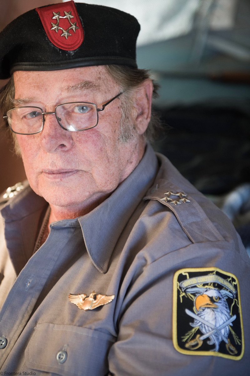 A close-up photograph of an older Caucasian man wearing a grey military-style uniform and a black beret with a red insignia. He wears glasses and has a patch on his left shoulder depicting an eagle and crossed swords.