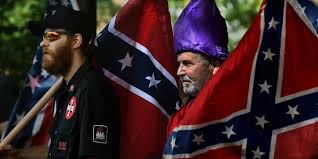 Two men at a white supremacist rally; one in a black shirt with KKK patches and a full beard, the other wearing a purple KKK hood and a grey beard, standing in front of Confederate flags.