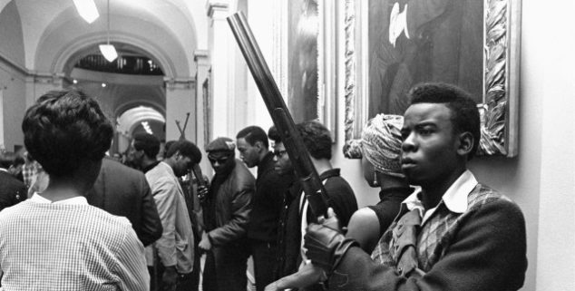 A black and white historical photograph of members of the Black Panther Party, some armed with rifles, standing in a hallway of a government building with paintings on the walls.