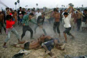A photograph showing a group of men in a rural Cambodian setting using long wooden poles to beat several individuals lying on the ground, while a crowd watches in the background.